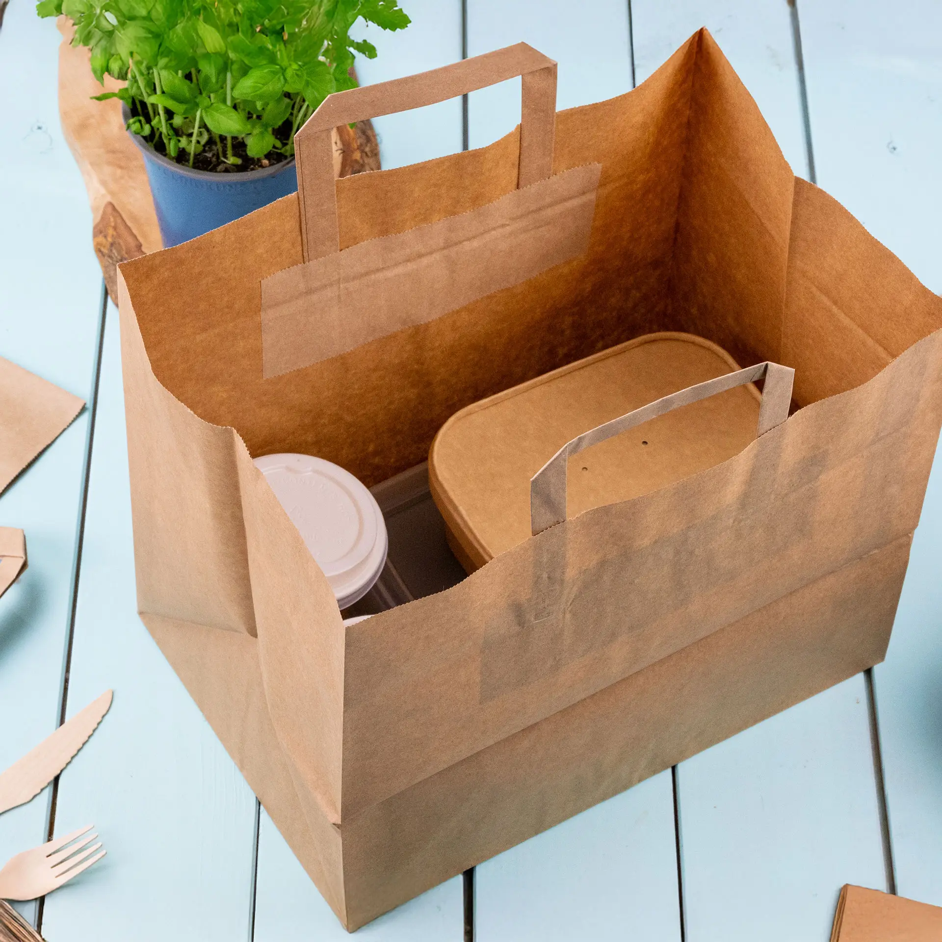 Brown paper carrier bag with flat handles; contains a food container and a cup, set against a light blue surface.